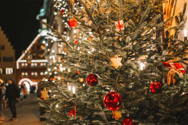 Outdoor Christmas tree at night time, decorated with baubles and lights, with lit up buildings in the background
